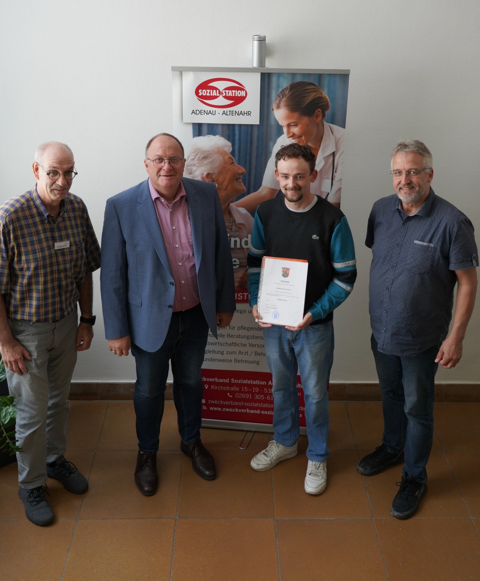 Vier Personen stehen nebeneinander und lächeln in die Kamera. Zweite Person von rechts ist Max Scherer, der seine Prüfung erfolgreich bestanden hat. Er hält eine Urkunde in der Hand. Neben ihm gratulieren drei Personen: links außen steht Fachbereichsleiter Bernhard Jüngling, daneben Verbandsvorsteher Guido Nisius und rechts außen der Leiter der Sozialstation Adenau-Altenahr Uwe Szymanski.