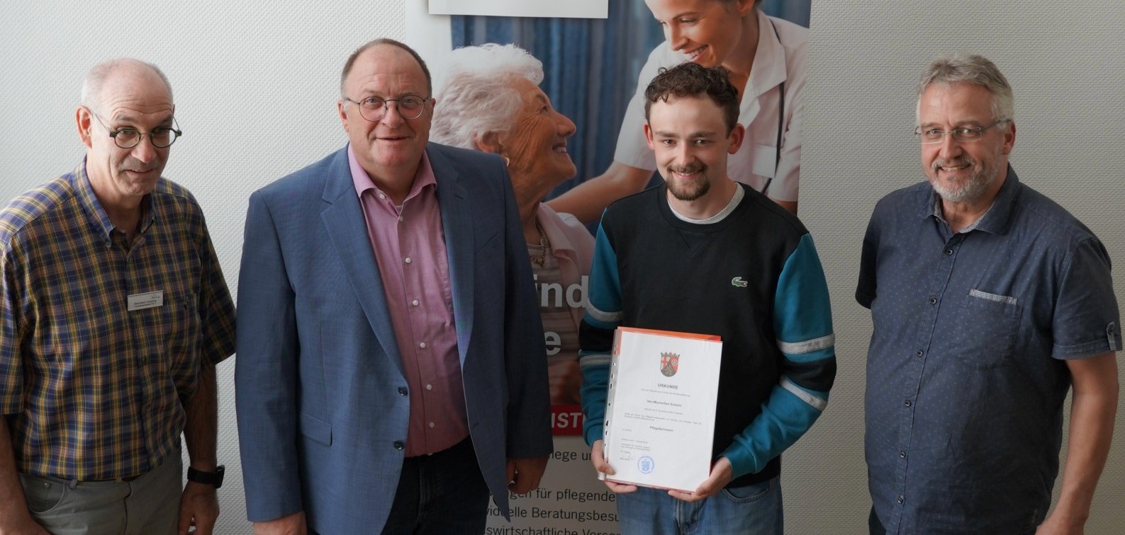 Vier Personen stehen nebeneinander und lächeln in die Kamera. Zweite Person von rechts ist Max Scherer, der seine Prüfung erfolgreich bestanden hat. Er hält eine Urkunde in der Hand. Neben ihm gratulieren drei Personen: links außen steht Fachbereichsleiter Bernhard Jüngling, daneben Verbandsvorsteher Guido Nisius und rechts außen der Leiter der Sozialstation Adenau-Altenahr Uwe Szymanski.