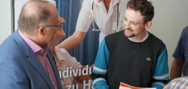 Max Scherer (r.) und Verbandsvorsteher Guido Nisius (l.) im Austausch über Zukunftspläne und Erfolge. Zwei Männer stehen nebeneinander und unterhalten sich freundlich. Links steht Verbandsvorsteher Guido Nisius, im blauen Anzug mit rosa Hemd, rechts Max Scherer, in dunkler Kleidung mit einem Dokument in der Hand. Beide wirken konzentriert und interessiert. Im Hintergrund hängt ein Poster mit einem Motiv aus der Pflege – darauf ist eine Pflegerin bei der Betreuung eines Patienten zu sehen, begleitet von teilweise lesbarem Text.