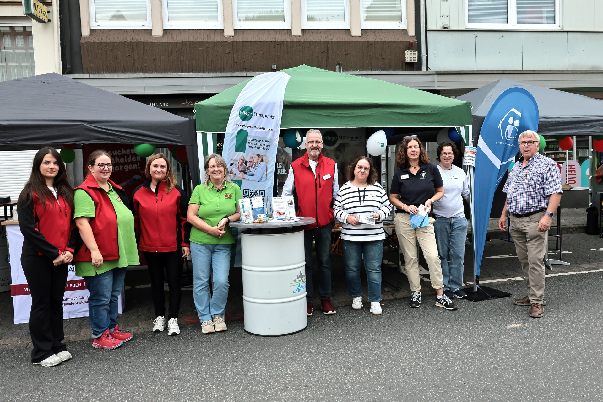 Gemeinsamer Informationsstand von Sozialstation, Tagespflege und Partnern beim Heimatfest Adenau 2026 Neun Personen stehen vor einem gemeinsamen Informationsstand auf dem Heimatfest in Adenau. Der Stand besteht aus mehreren Pavillons mit Informationsmaterialien, Flyern und Roll-ups der Sozialstation Adenau-Altenahr, des Pflegestützpunkts, der Tagespflege, der Gemeindeschwesterplus und des Fördervereins. Die Beteiligten präsentieren gemeinsam ihre Angebote rund um Pflege, Betreuung, Beratung und Unterstützung im Alter. Im Hintergrund sind weitere Stände sowie Gebäude in der Adenauer Innenstadt zu sehen.