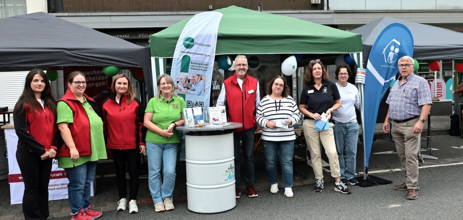 Gemeinsamer Informationsstand von Sozialstation, Tagespflege und Partnern beim Heimatfest Adenau 2026 Neun Personen stehen vor einem gemeinsamen Informationsstand auf dem Heimatfest in Adenau. Der Stand besteht aus mehreren Pavillons mit Informationsmaterialien, Flyern und Roll-ups der Sozialstation Adenau-Altenahr, des Pflegestützpunkts, der Tagespflege, der Gemeindeschwesterplus und des Fördervereins. Die Beteiligten präsentieren gemeinsam ihre Angebote rund um Pflege, Betreuung, Beratung und Unterstützung im Alter. Im Hintergrund sind weitere Stände sowie Gebäude in der Adenauer Innenstadt zu sehen.