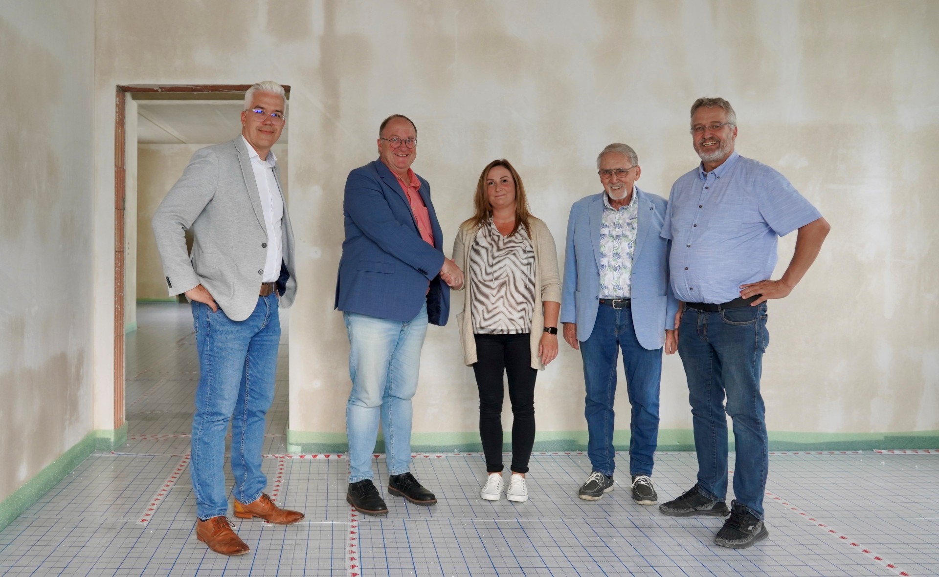 Fünf Personen stehen in einem Raum mit unverputzten Wänden und einem Boden, der mit einem rot-weiß markierten Raster versehen ist. Die Gruppe besteht aus vier Männern und einer Frau. Von links nach rechts sind zu sehen: Dominik Gieler (stellvertretender Verbandsvorsteher), Guido Nisius (Verbandsvorsteher), Martina Romes (zukünftige Leitung der Tagespflege in Liers), Jürgen de Temple (stellvertretender Verbandsvorsteher) und Uwe Szymanski (Leiter der Sozialstation).