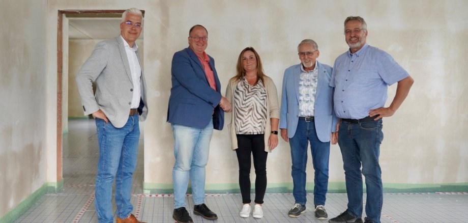 Fünf Personen stehen in einem Raum mit unverputzten Wänden und einem Boden, der mit einem rot-weiß markierten Raster versehen ist. Die Gruppe besteht aus vier Männern und einer Frau. Von links nach rechts sind zu sehen: Dominik Gieler (stellvertretender Verbandsvorsteher), Guido Nisius (Verbandsvorsteher), Martina Romes (zukünftige Leitung der Tagespflege in Liers), Jürgen de Temple (stellvertretender Verbandsvorsteher) und Uwe Szymanski (Leiter der Sozialstation).